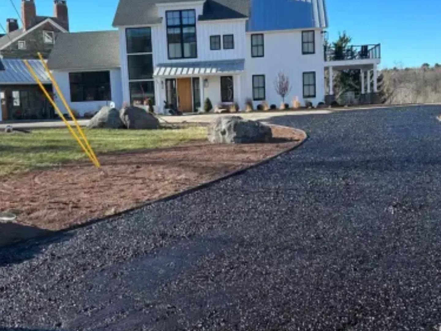 Freshly laid dark chip seal driveway curving past a lawn to a white farmhouse