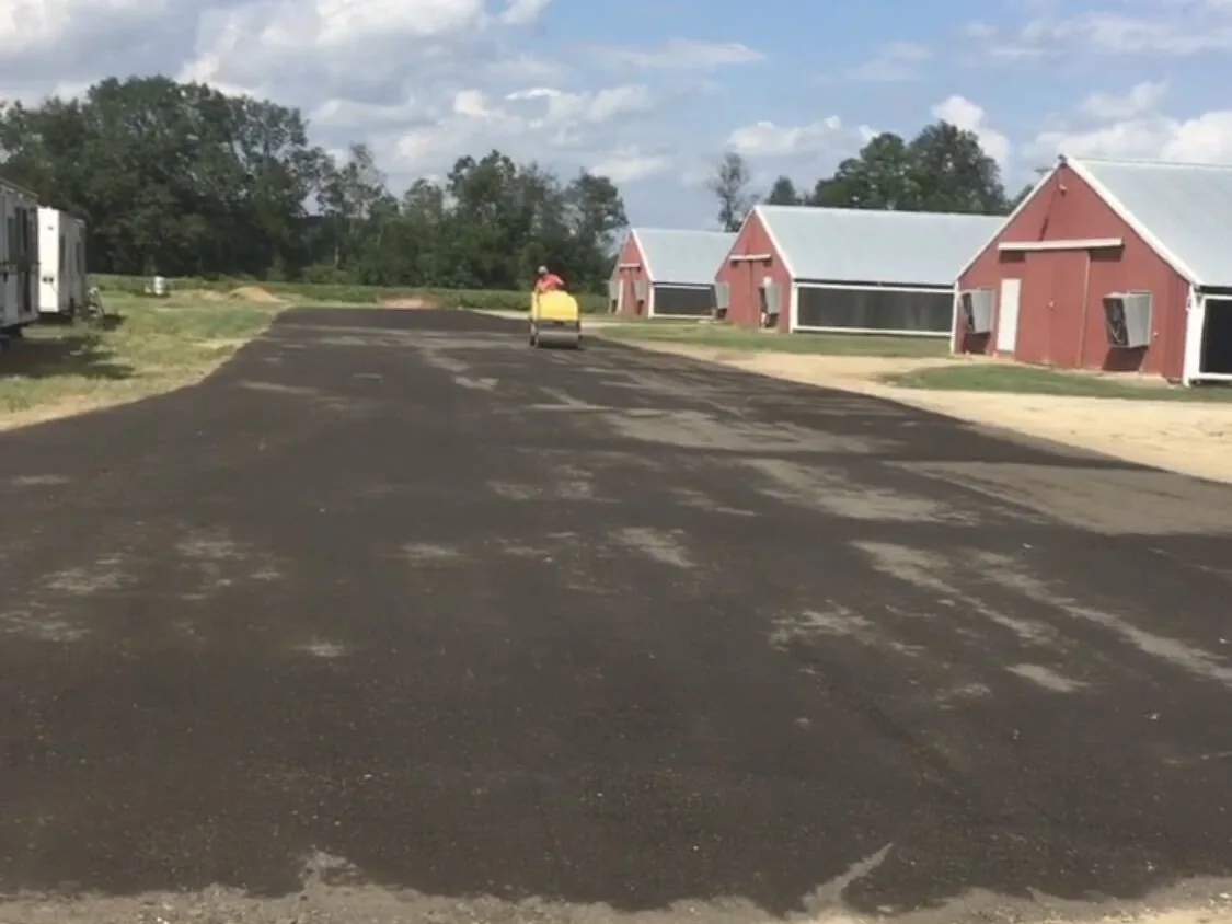Roller compacting a wide paved lot beside red farm buildings, parking lot paving