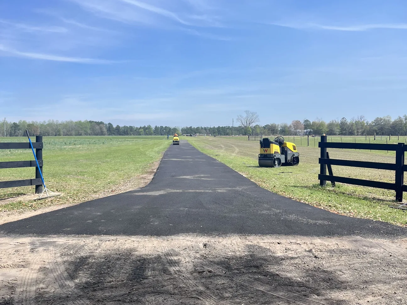 Long freshly paved private drive through a fenced pasture with rollers at work, private road paving