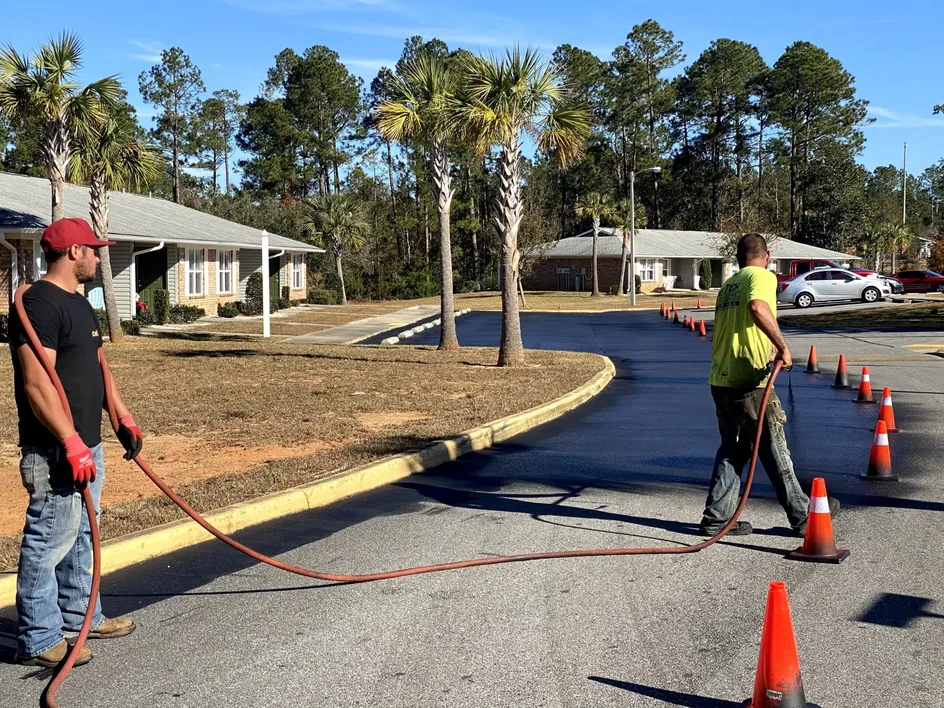Two man crew spraying sealer on an asphalt drive lined with traffic cones, sealcoating