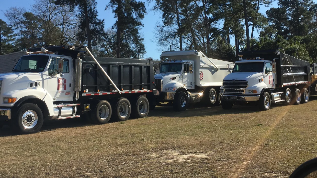 Three tri axle dump trucks lined up under pines, asphalt paving