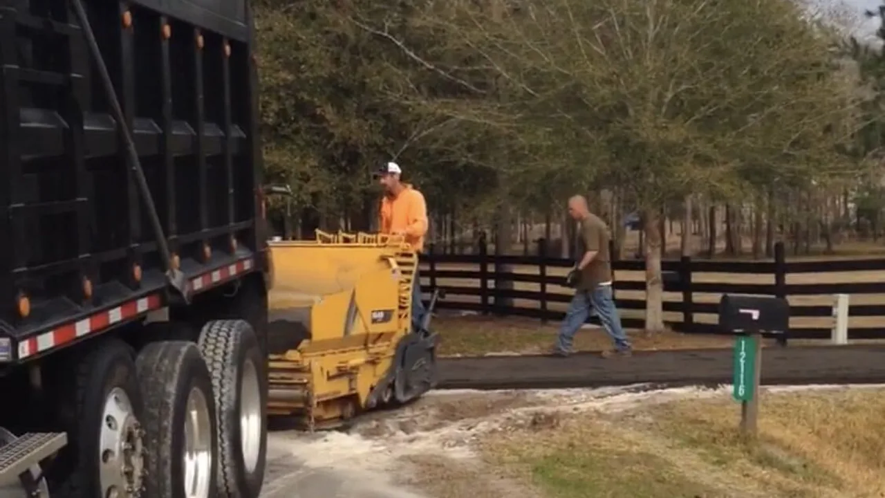 Dump truck feeding a paving machine as the crew lays a new drive along a fence